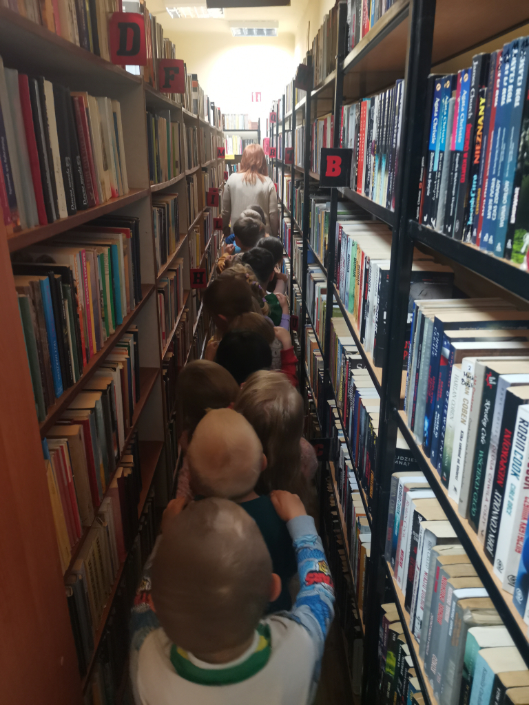 Zdjęcie przedstawia grupę przedszkolaków zwiedzających bibliotekę bawiąc się w zabawę Wesoły pociąg